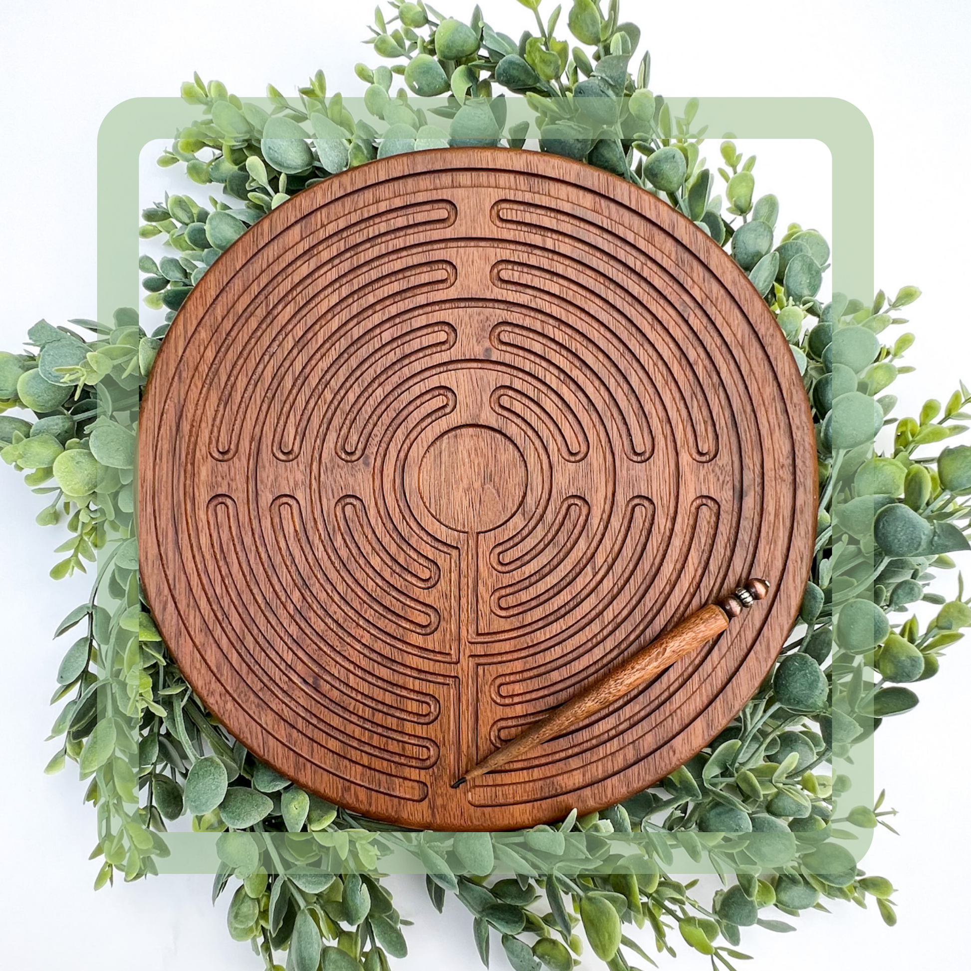 Handcrafted wooden labyrinth with a stylus, placed on a bed of artificial green leaves. The labyrinth has a circular design with multiple grooves, and is carved from walnut wood.
